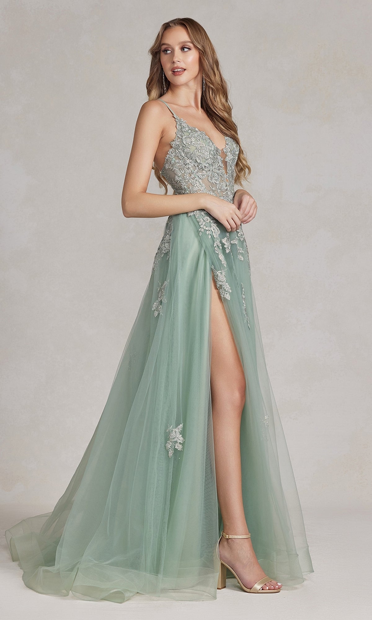 A woman stands against a neutral background wearing the Open-Back Long Prom Ball Gown with Beads, a light green, floor-length A-line dress featuring floral embellishments, beaded lace appliques, a deep V-neckline, thin straps, and a high slit.